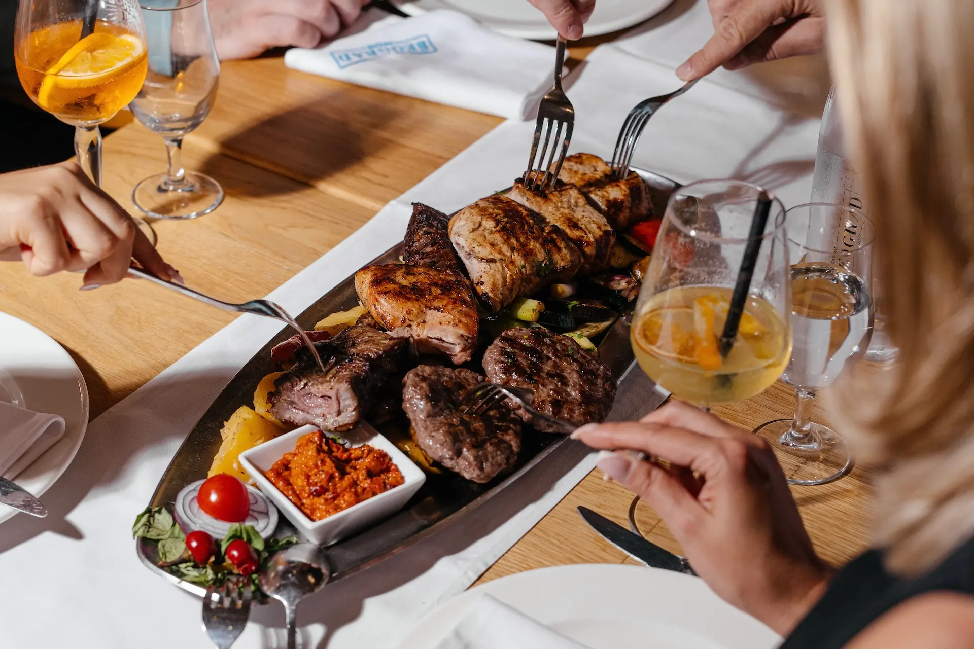 Eine Gruppe von Menschen teilt sich an einem Holztisch eine Platte mit gegrilltem Fleisch, Gemüse und Dip-Soße, daneben stehen Getränke und weiße Servietten.