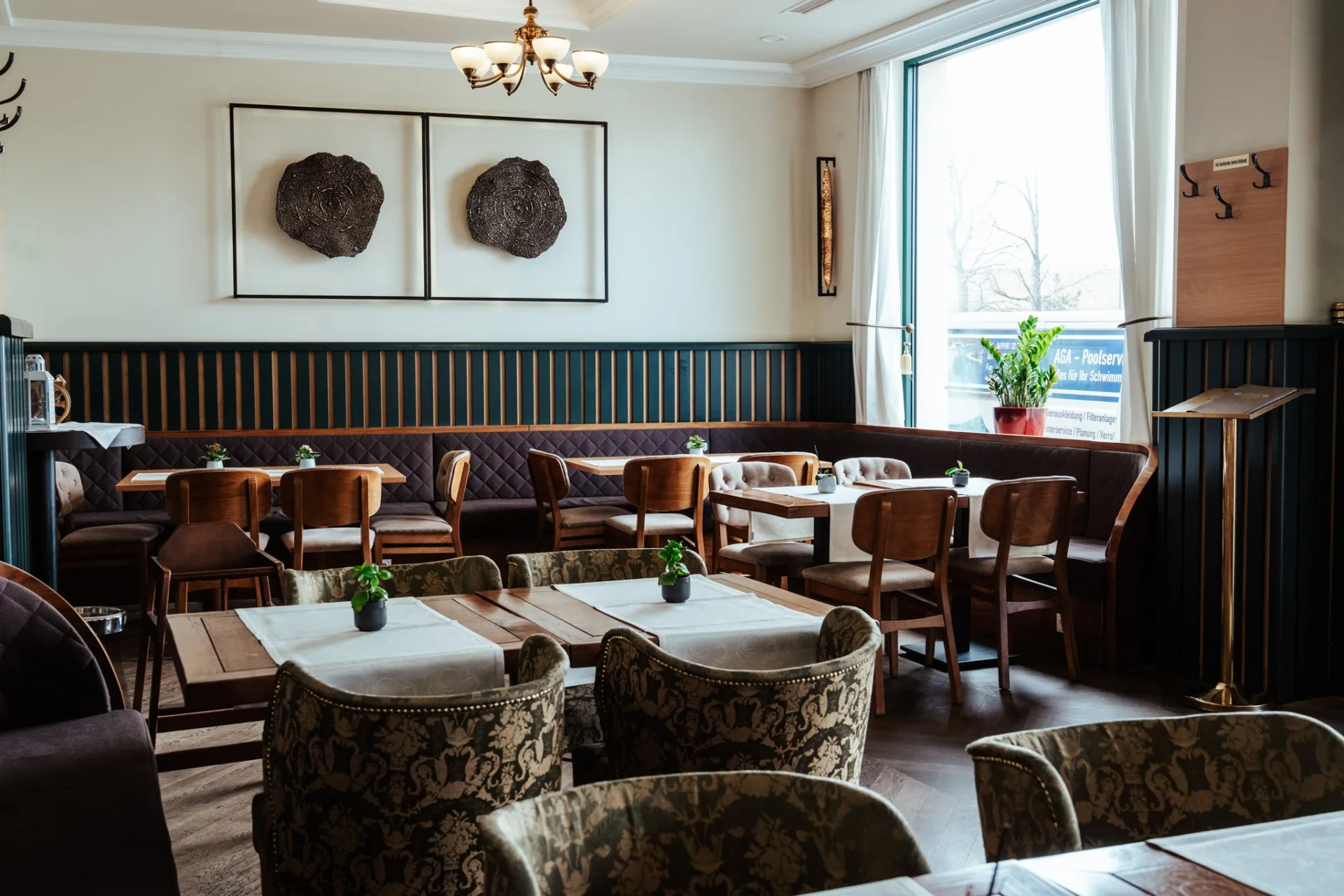 Ein gemütliches, modernes Café mit Holztischen, gepolsterten Stühlen, einer langen gepolsterten Bank, Topfpflanzen auf den Tischen, einem großen Fenster, Wandkunst und eleganter Beleuchtung.