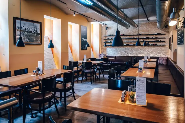Ein modernes, gemütliches Restaurant mit Holztischen und -stühlen, Hängelampen, Wandkunst und Regalen. Auf den Tischen stehen Speisekarten und kleine Pflanzen, und durch die hohen Fenster strömt Sonnenlicht herein. Der Raum ist menschenleer.