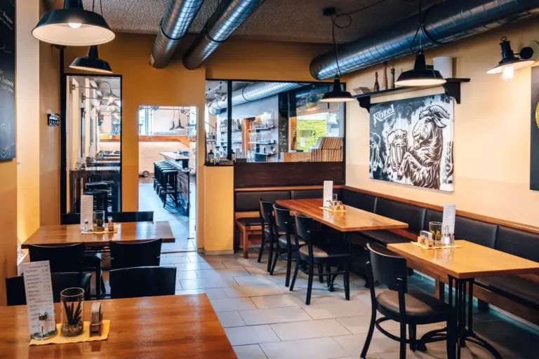Ein gemütliches, leeres Restaurant mit Holztischen und schwarzen Stühlen, Deckenlüftern im Industriestil, warmen gelben Wänden und einem schwarz-weißen Wandgemälde mit einem Hahn an einer Wand. Speisekarten, Salz- und Pfefferstreuer stehen auf den Tischen.