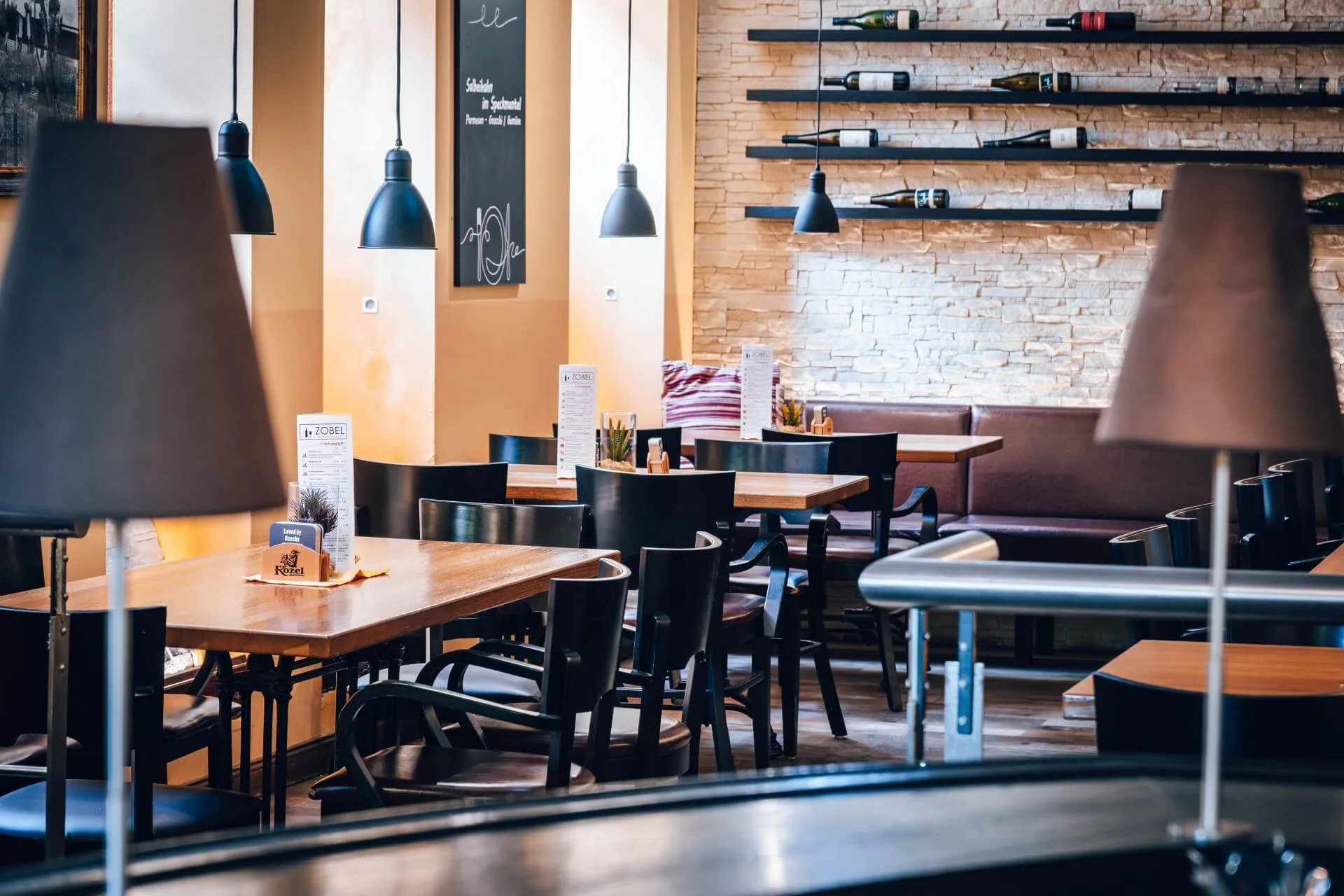 Ein modernes Restaurant mit Holztischen und schwarzen Stühlen, von der Decke hängenden Pendelleuchten und dekorativen Weinflaschen in Regalen entlang einer weißen Backsteinwand. Der Raum wirkt leer und einladend.