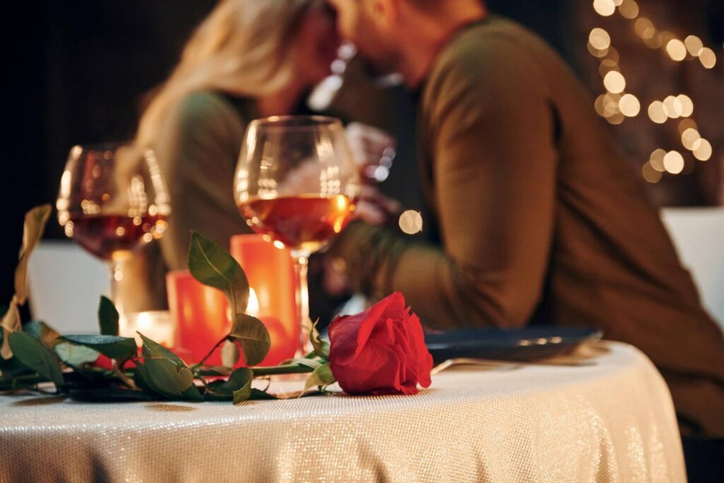 Ein romantisch gedeckter Tisch mit zwei Weingläsern, Kerzen und einer roten Rose im Mittelpunkt. Im unscharfen Hintergrund sitzt ein Paar dicht beieinander und erinnert an die intime Atmosphäre in den besten Romantischen Restaurants in Wien.