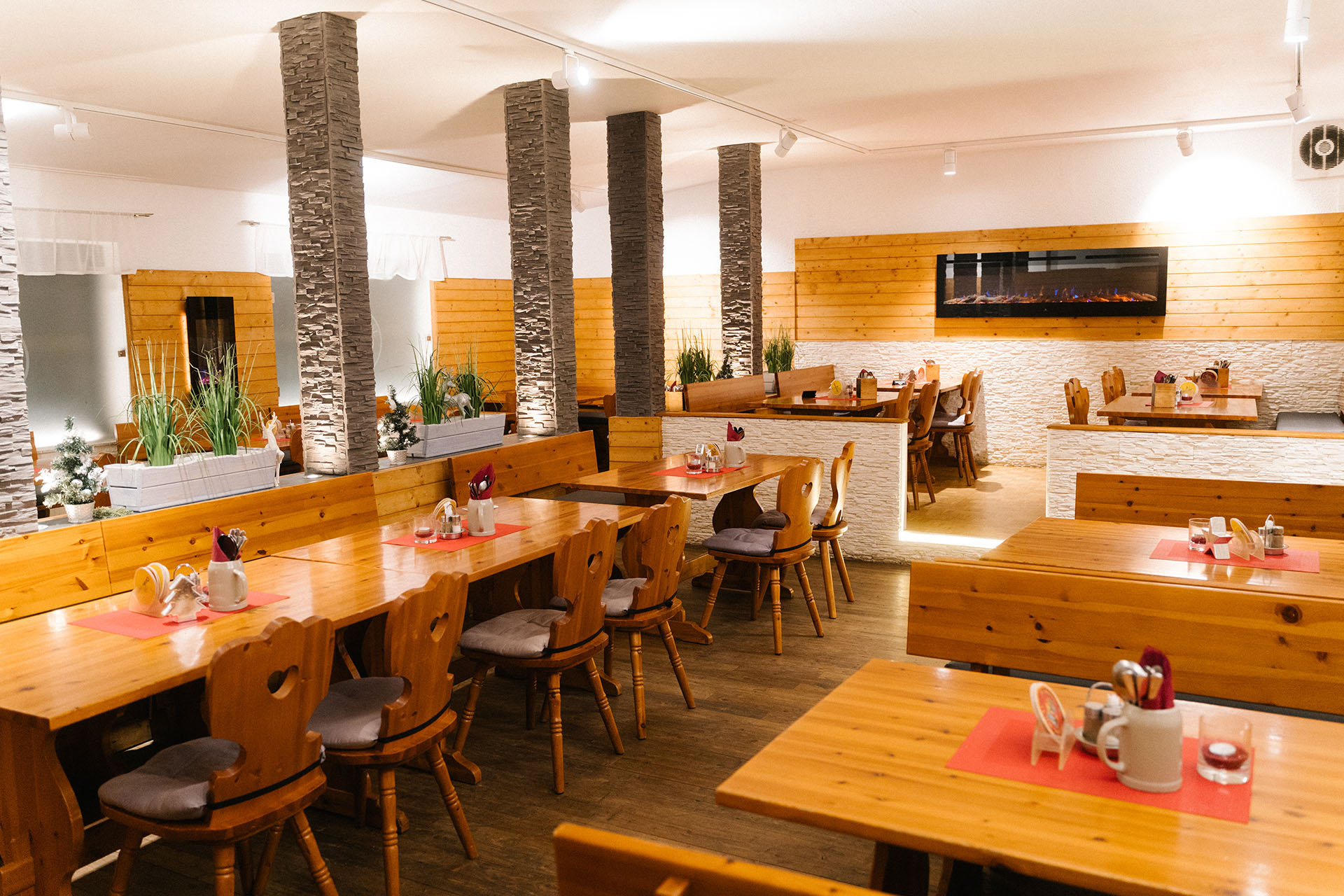 Ein gemütliches Restaurant mit Holztischen und -stühlen, Steinsäulen und Topfpflanzen. Der Raum ist warm beleuchtet, mit einem modernen elektrischen Kamin an der Wand und roten Tischsets auf jedem Tisch.