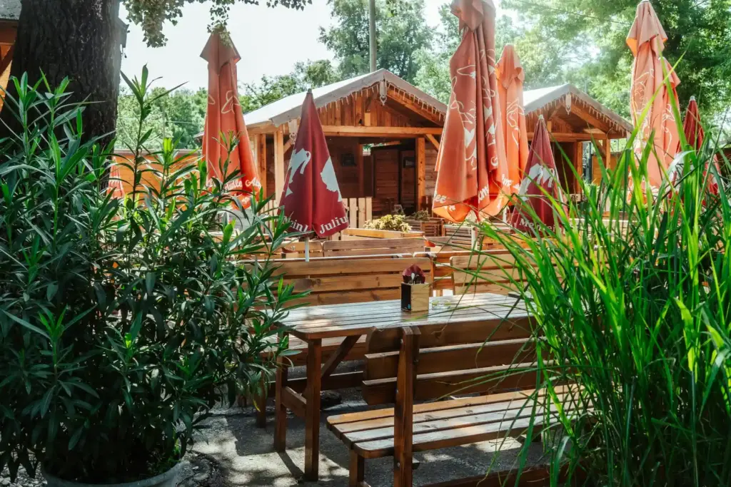 Picknicktische und -bänke aus Holz stehen draußen zwischen grünen Pflanzen und hohem Gras, große orangefarbene Sonnenschirme spenden Schatten. Kleine Holzhütten stehen an einem sonnigen Tag im Hintergrund unter Bäumen.