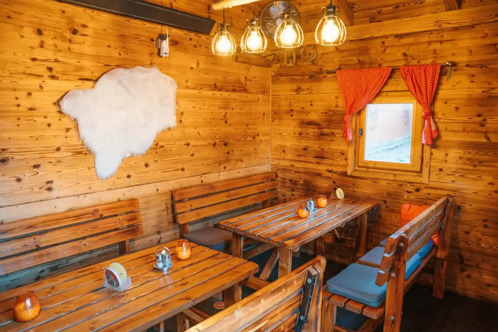 Ein gemütlicher Holzraum mit Bänken und Tischen, warmes Licht von Hängelampen, ein kleines Fenster mit einem roten Vorhang und eine weiße Felldekoration an der Wand schaffen eine rustikale und einladende Atmosphäre.