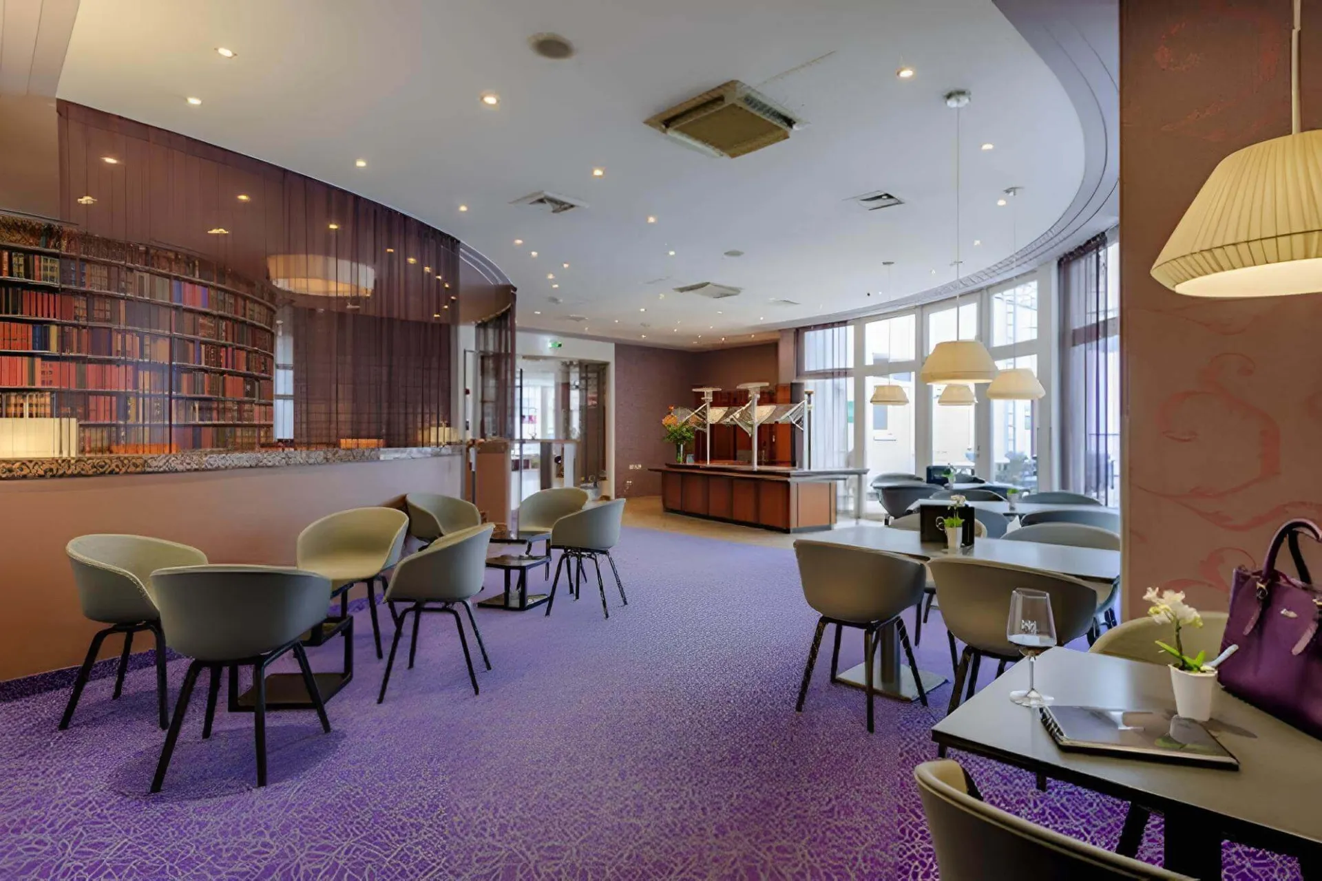 Eine moderne Lounge mit lila Teppich, grauen Stühlen und kleinen Tischen. Auf der linken Seite befindet sich eine Bar mit Bücherregalen, während große Fenster und Hängelampen den Raum erhellen. Auf einigen Tischen stehen Getränke und eine Handtasche.