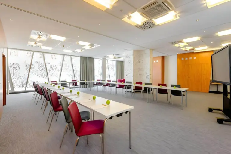 Ein moderner Konferenzraum mit U-förmig angeordneten Tischen, an denen jeweils ein roter Stuhl und ein grüner Apfel auf dem Tisch stehen. Große Fenster lassen natürliches Licht herein, und in einer Ecke steht eine Projektionsfläche.