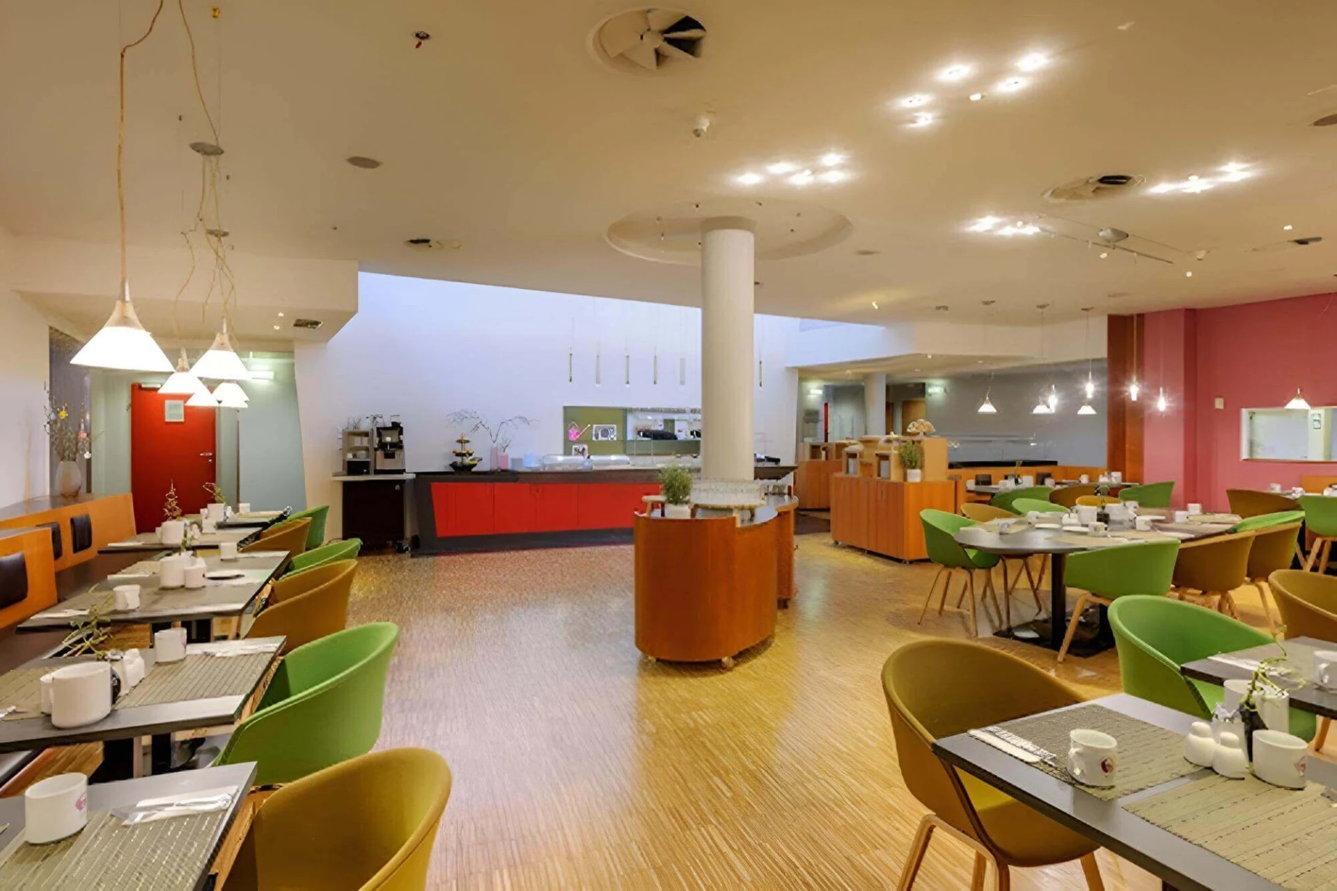 Moderne Cafeteria mit hellen Holzböden, gedeckten Tischen, grünen und braunen Stühlen, Hängelampen und einem Thekenbereich im Hintergrund mit Kaffeemaschinen und Essensstationen. Der Raum ist hell beleuchtet und einladend.