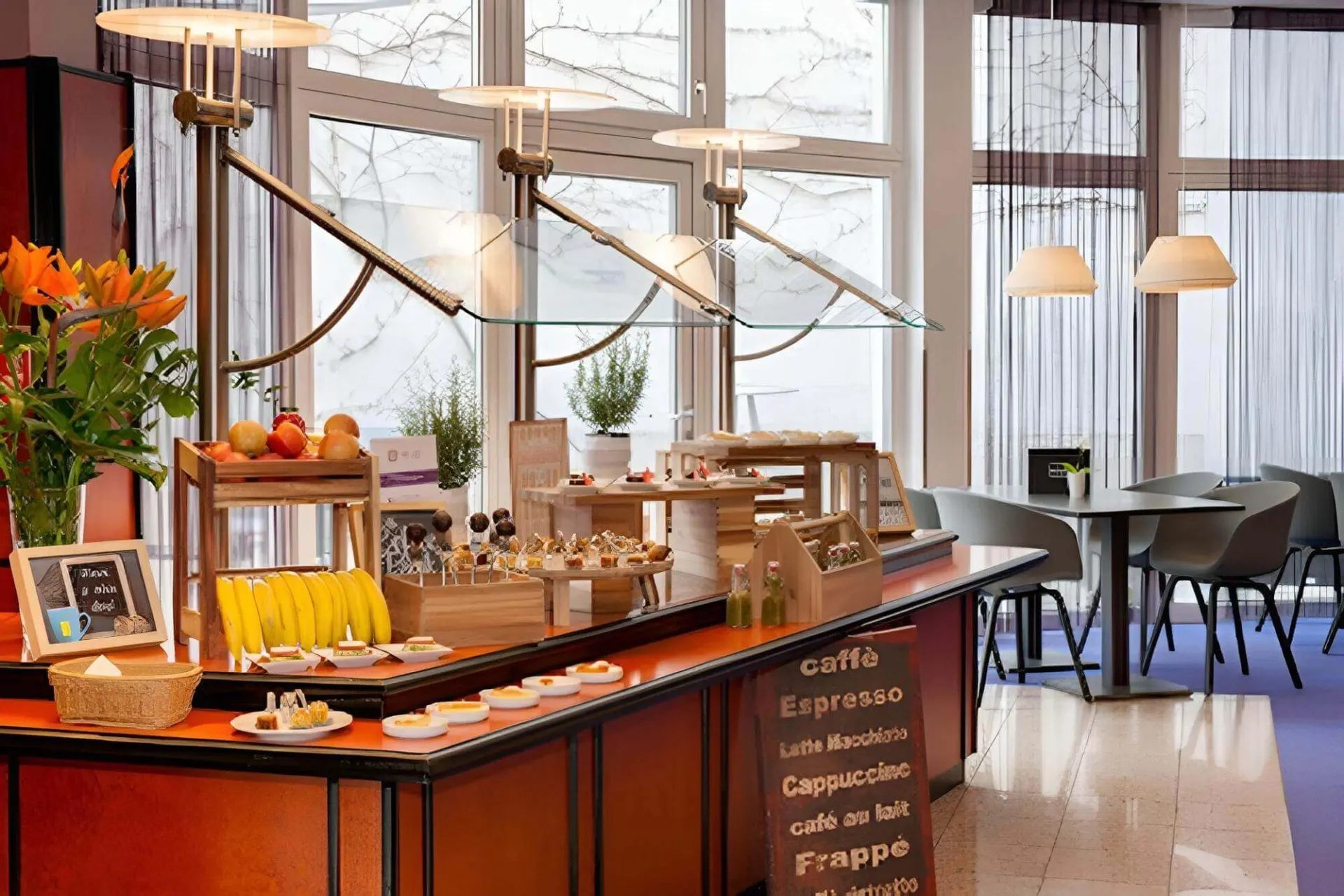 Ein Frühstücksbuffet mit Gebäck, Obst und Getränken steht auf einer Holztheke in einem hellen Essbereich mit großen Fenstern, Pflanzen und Tischen mit modernen Stühlen im Hintergrund.