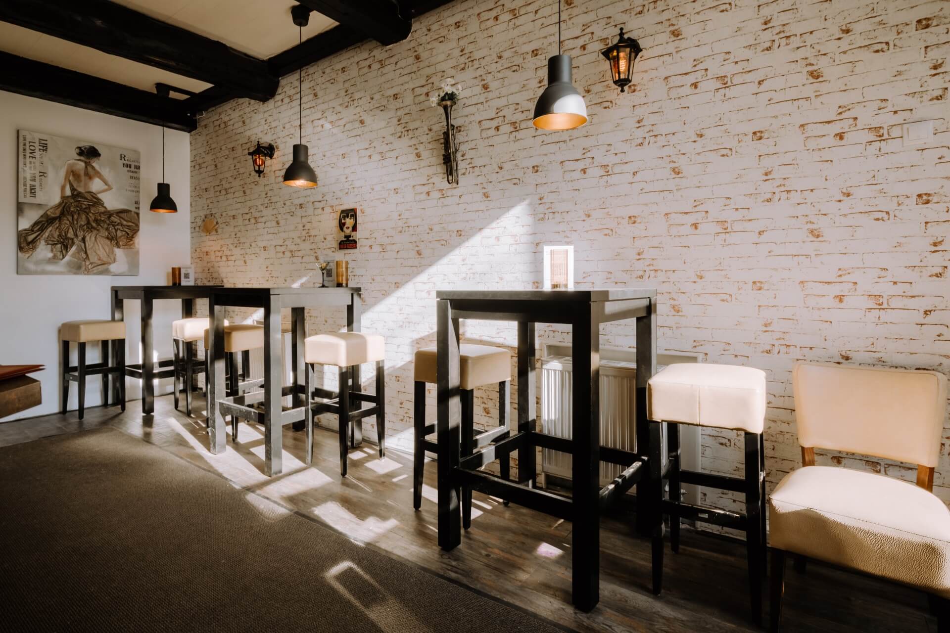 Ein gemütliches Café mit hohen Tischen und cremefarbenen Hockern vor einer weißen Backsteinwand, dekoriert mit modernen Hängelampen und gerahmter Kunst, die von einem nahen Fenster beleuchtet wird.