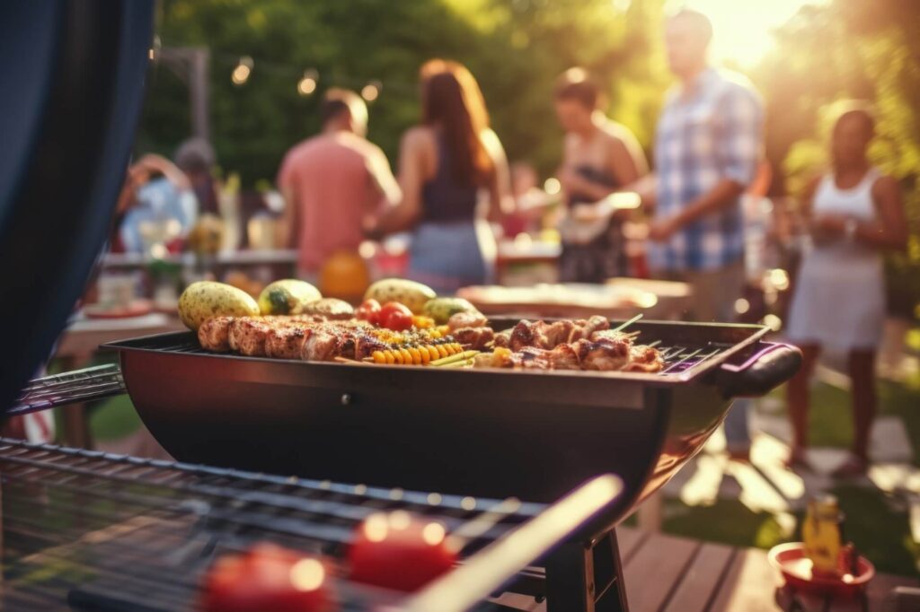 Eine Nahaufnahme einer Grillszene mit Spießen, Mais und Gemüse, die auf dem Grill brutzeln, während sich im unscharfen Hintergrund Menschen bei einem Treffen im Freien an einem sonnigen Tag unterhalten.