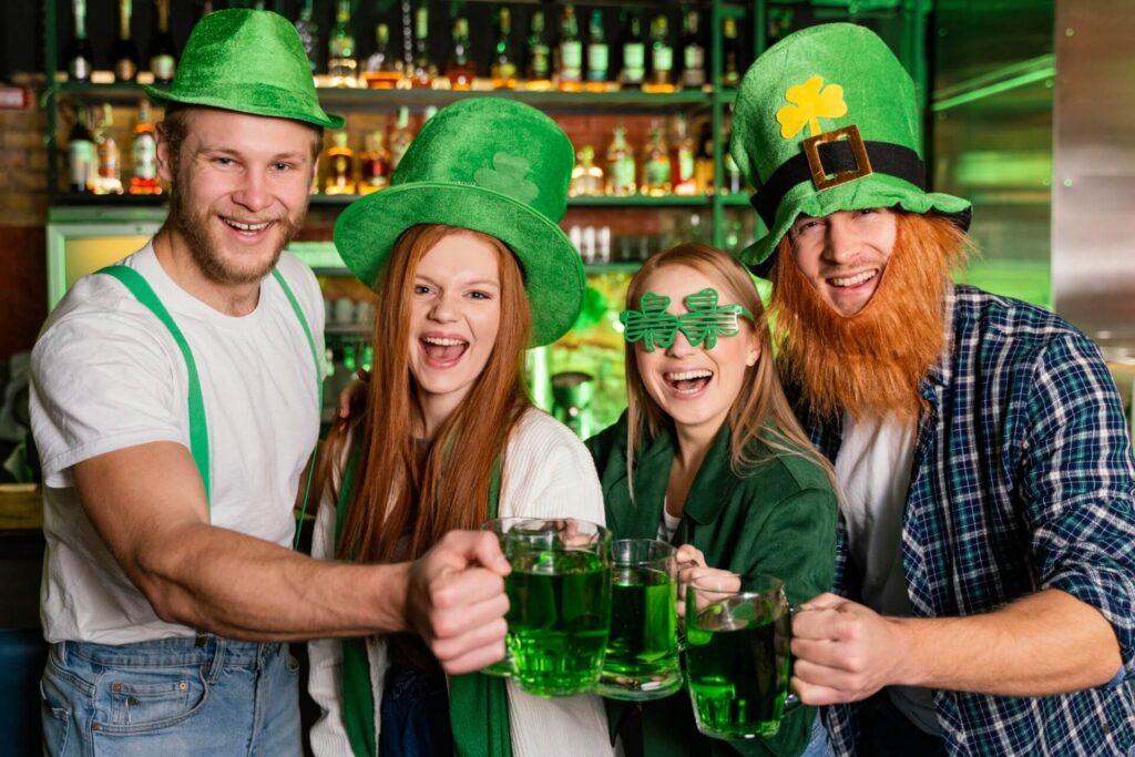 Vier Personen in grüner Saint-Patricks-Day-Kleidung, einschließlich Hüten und lustigen Gläsern, stoßen in einer festlichen Bar fröhlich mit grünen Bierkrügen an.