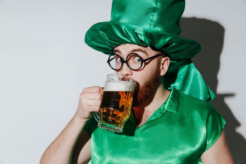 Ein Mann mit großem grünen Hut, runder Brille und grünem Hemd trinkt Bier aus einem Glasbecher. Das festliche Kostüm eignet sich perfekt für die Feier des Saint Patricks Day.