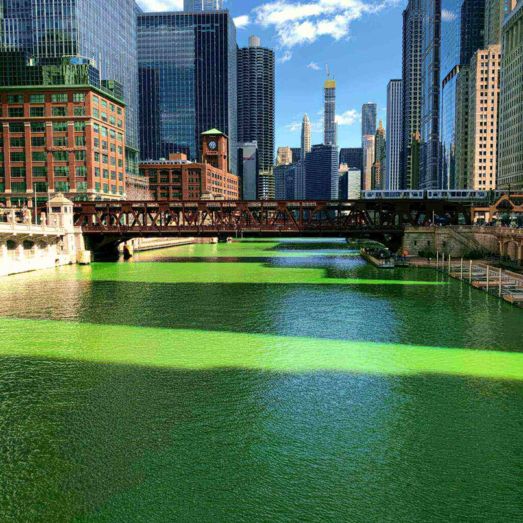 Ein für den Saint Patricks Day hellgrün gefärbter Stadtfluss fließt unter einer Metallbrücke mit einem Zug hindurch; hohe moderne Wolkenkratzer und historische Backsteinbauten säumen das Wasser unter einem blauen Himmel mit vereinzelten Wolken.