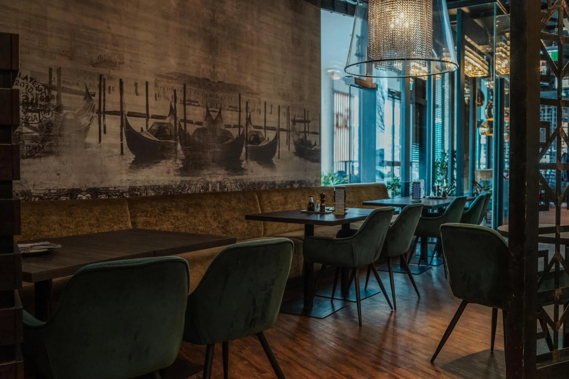 Ein modernes Restaurant mit grünen Samtstühlen, dunklen Holztischen, gedämpfter Beleuchtung und einem großen Wandgemälde mit Gondeln auf dem Wasser, das eine gemütliche und elegante Atmosphäre schafft.