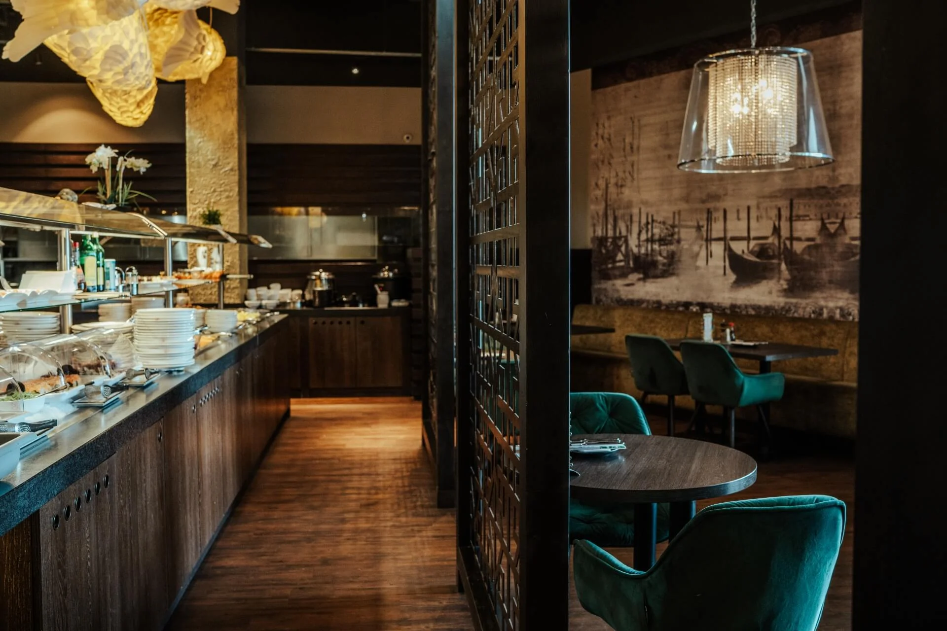 Ein gemütliches Restaurant mit Holzböden, grünen Samtstühlen, einem runden Tisch, einer Buffet-Theke mit Geschirr und warmer Beleuchtung. Ein großes Wandkunstwerk und dekorative Hängelampen tragen zum Ambiente bei.