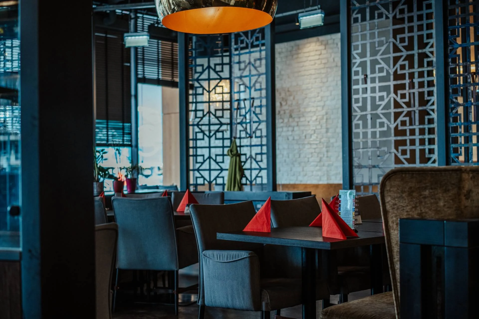 Modernes Restaurant mit zeitgenössischer Einrichtung, Holztischen, gepolsterten Stühlen, geometrischen Wandpaneelen und roten Servietten, die ordentlich auf den Tischen gefaltet sind. Die warme Beleuchtung schafft eine gemütliche Atmosphäre.