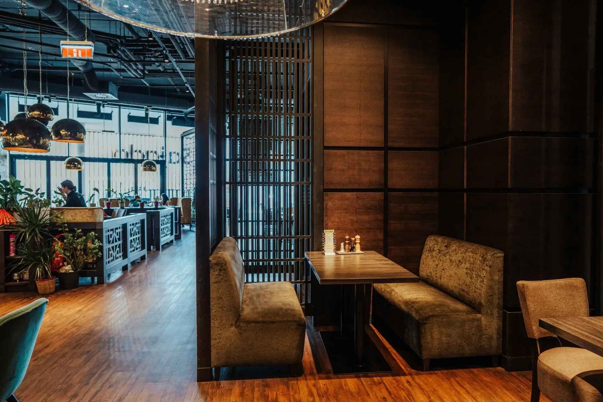 Ein modernes Restaurant mit Holzböden, gemütlichen Kabinen mit grün gepolsterten Sitzen, gedämpfter Beleuchtung und dekorativen Pflanzen. Die Atmosphäre ist warm und einladend, mit natürlichem Licht, das durch große Fenster einfällt.