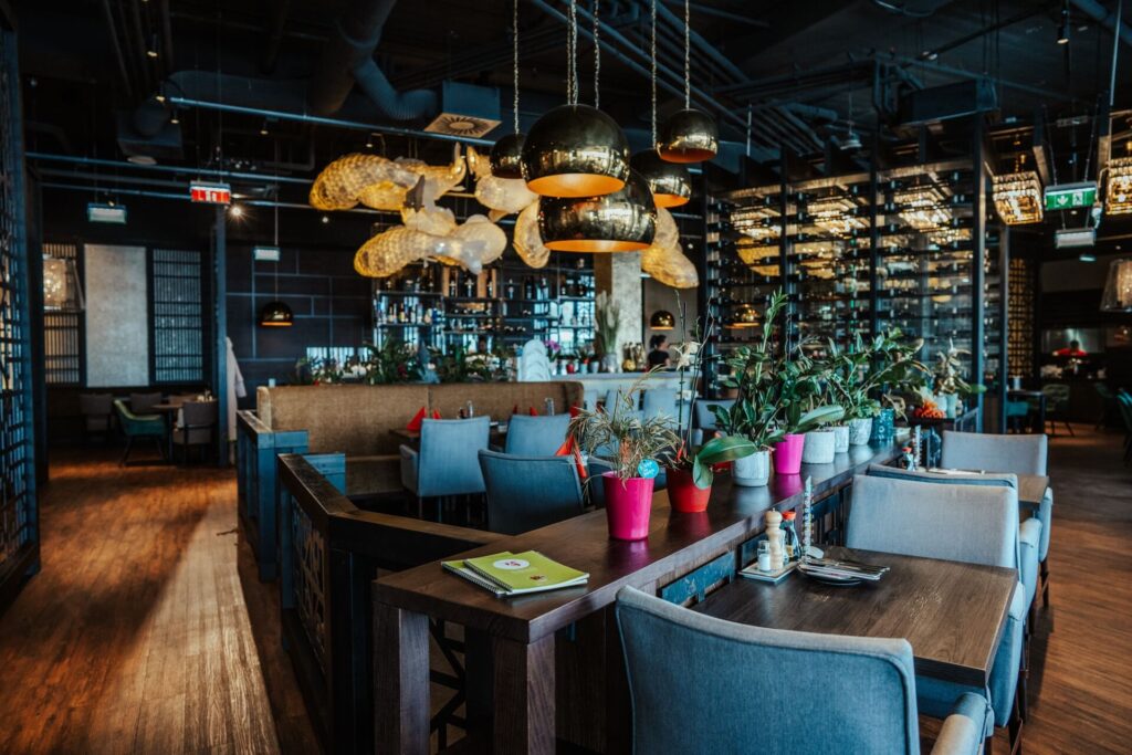 Ein modernes Restaurant mit Holzböden, blaugrauen Stühlen, Topfpflanzen auf den Tischen, goldenen Pendelleuchten und großen Fischskulpturen, die von der Decke hängen. Im Hintergrund sind Regale und eine Bar zu sehen.
