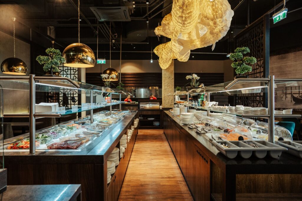 Ein modernes Restaurantbuffet mit Reihen von Tabletts, Salaten und Desserts unter Glasabdeckungen. Warme Beleuchtung, goldene Lampen und dekorative Hängeelemente schaffen eine elegante Essatmosphäre.
