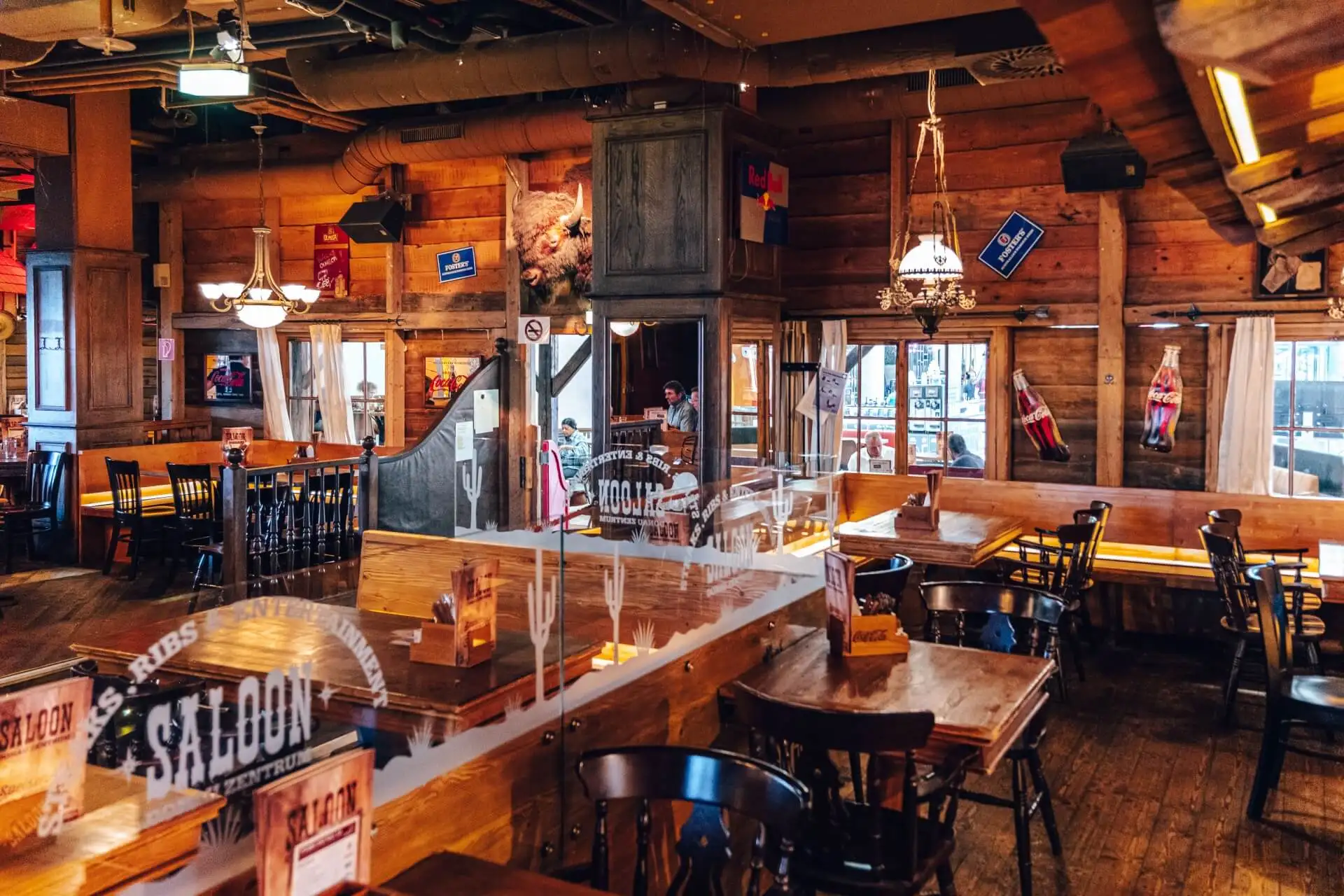 Ein rustikales Restaurant im Western-Stil mit Holztischen und -stühlen, warmer Beleuchtung, Vintage-Dekor, Flaggen und großen Fenstern, die viel Tageslicht hereinlassen. Der Raum wirkt gemütlich und einladend, mit einer ungezwungenen Essatmosphäre.