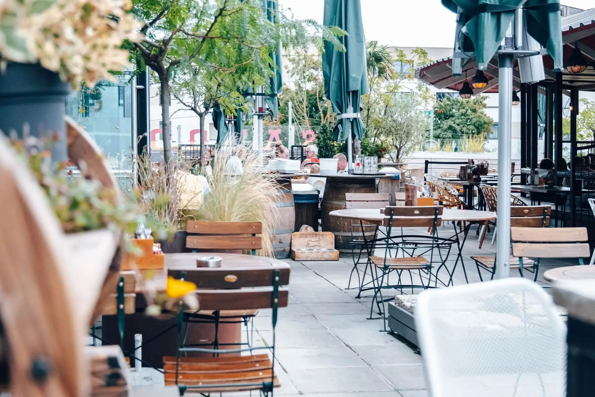 Restaurantterrasse im Freien mit leeren Holztischen und -stühlen, Grünzeug und Topfpflanzen sowie großen Sonnenschirmen. Der Bereich wirkt entspannt und einladend, mit sanftem Tageslicht und ein paar Menschen im Hintergrund.