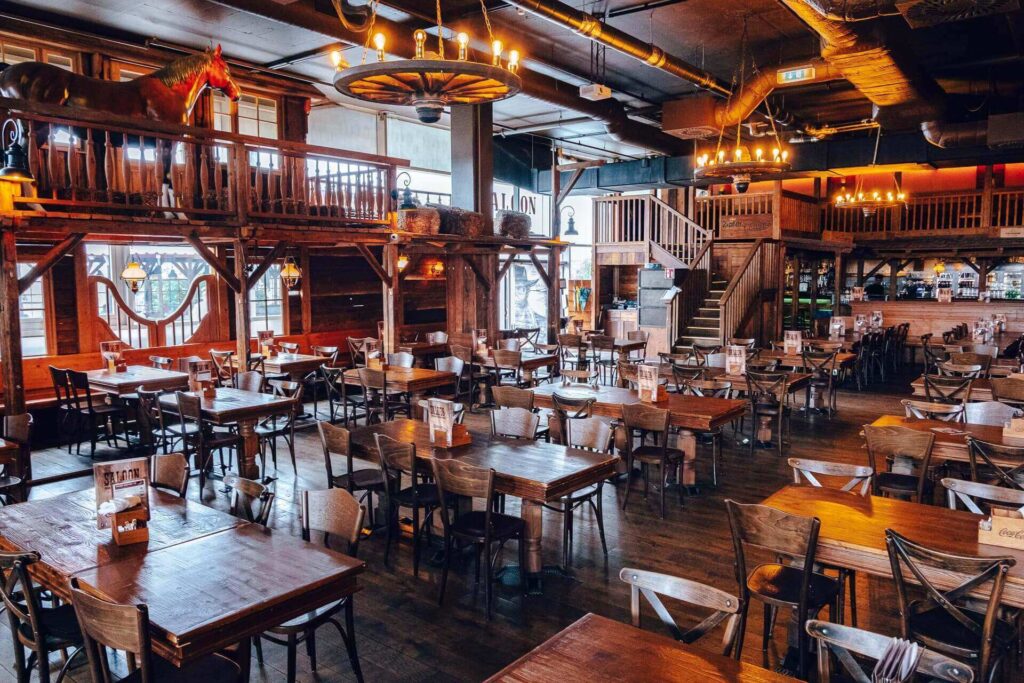 Ein geräumiges Western-Restaurant in Wien mit Holztischen und -stühlen, rustikalem Dekor, Wagenrad-Lüstern, einem Balkon mit einer Pferdestatue und einem Barbereich. Eine außergewöhnliche Location in Wien - perfekt für einzigartige Zusammenkünfte.