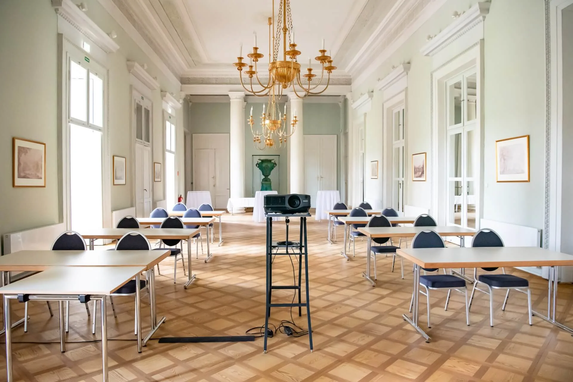 Ein geräumiger, eleganter Konferenzraum mit Kronleuchtern, Parkettfußboden und großen Fenstern. Mehrere Tische und Stühle sind in Reihen angeordnet, vor denen ein Projektor auf einem Ständer steht.