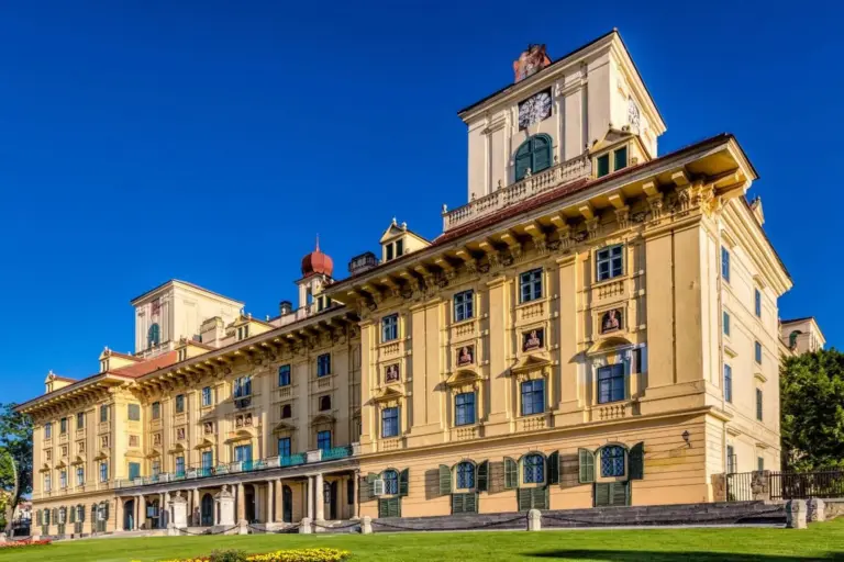Ein prächtiges, historisches, gelbes Gebäude mit verschnörkelten architektonischen Details, vielen Fenstern und Balkonen vor einem klaren blauen Himmel mit grünem Gras und Blumen im Vordergrund.