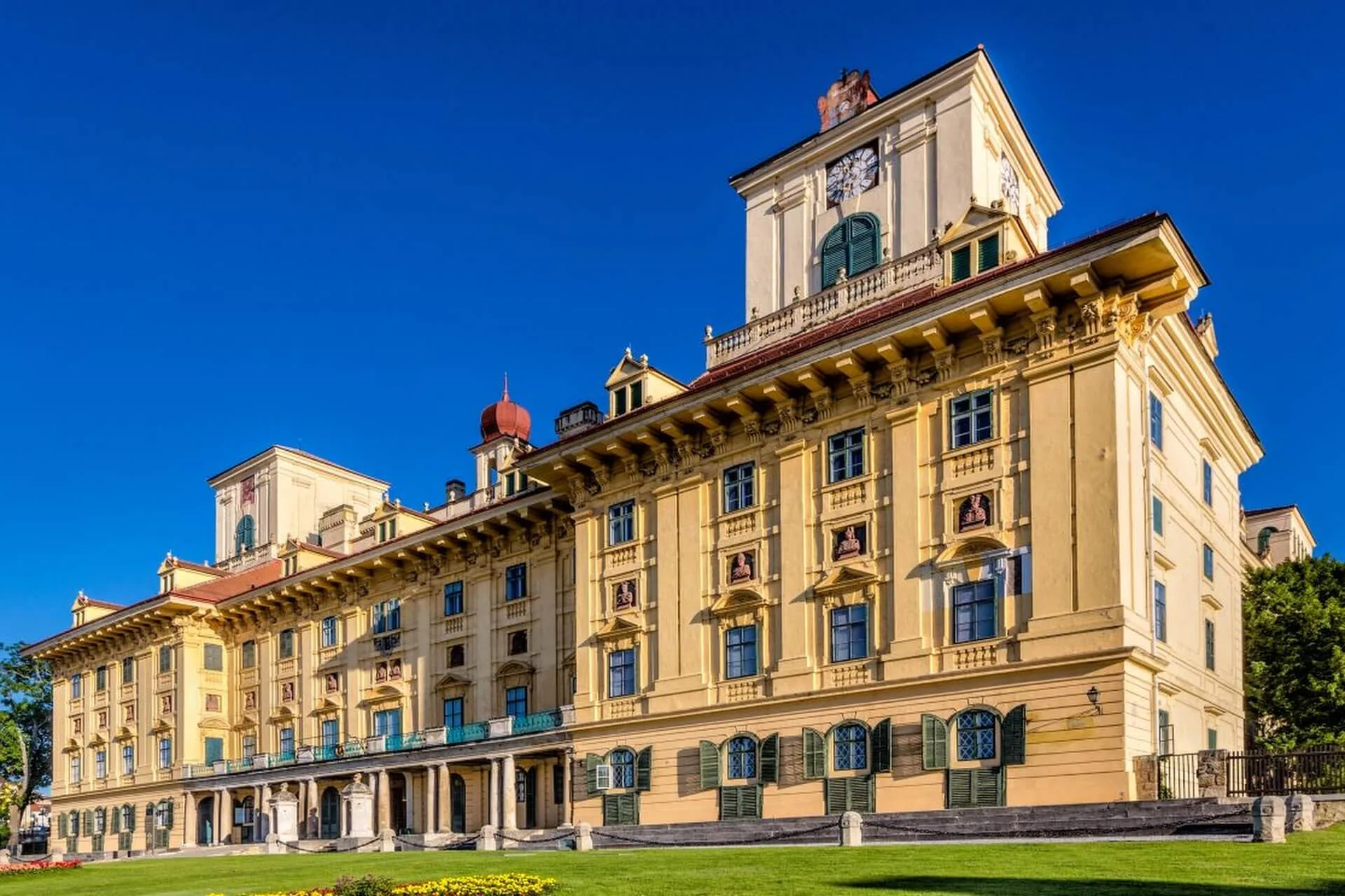 Ein prächtiges, historisches, gelbes Gebäude mit verschnörkelten architektonischen Details, vielen Fenstern und Balkonen vor einem klaren blauen Himmel mit grünem Gras und Blumen im Vordergrund.