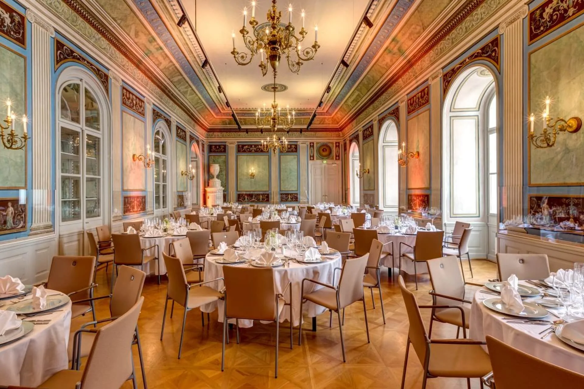 Eleganter Bankettsaal mit verzierter Decke, Kronleuchtern, großen Fenstern und runden Tischen mit weißen Tischtüchern, Gläsern und Servietten, umgeben von braunen Stühlen. Die Wände sind mit klassischen Kunstwerken und Stuck verziert.