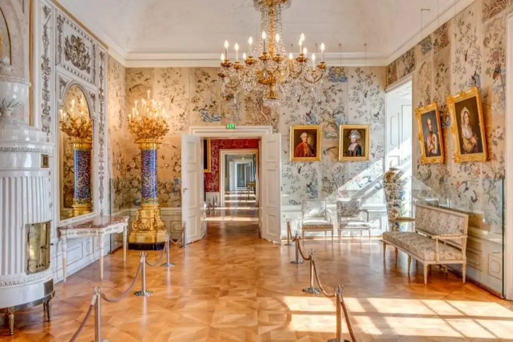 Ein prächtiger, verschnörkelter Palastflur mit Kronleuchtern, gemusterten Tapeten, vergoldeten Spiegeln, Porträtgemälden, eleganten Möbeln und polierten Holzböden, die zum Schutz durch Seile getrennt sind.