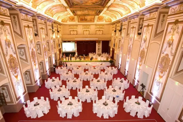 Ein prächtiger, reich verzierter Bankettsaal mit hohen, verzierten Decken, eleganten Wandgestaltungen und rotem Teppich. Zahlreiche runde Tische mit weißen Tüchern und blumigen Tafelaufsätzen sind fein säuberlich für eine Veranstaltung angeordnet. Am hinteren Ende ist eine Bühne aufgebaut.