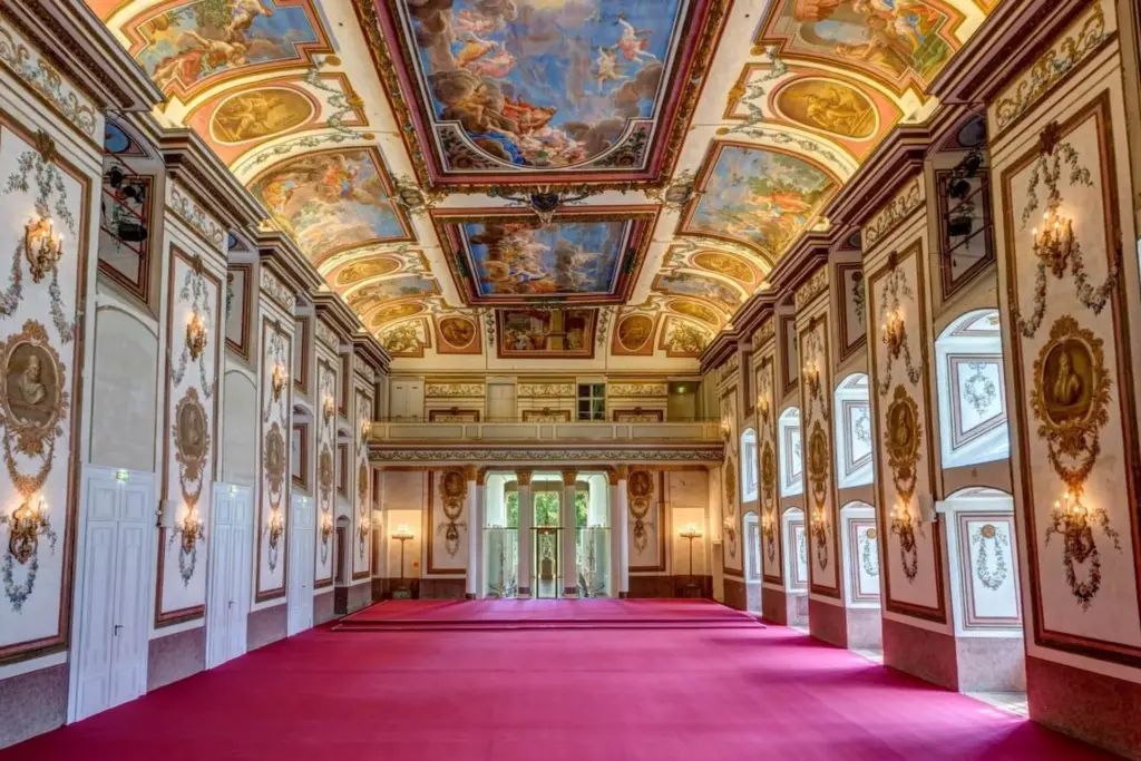 Ein prächtiger, verzierter Saal mit einer hohen, bemalten Decke, detaillierten gold-weißen Wänden, großen Bogenfenstern und einem großen roten Teppich auf dem Boden. Der Saal ist reich verziert und mit natürlichem Licht erfüllt.
