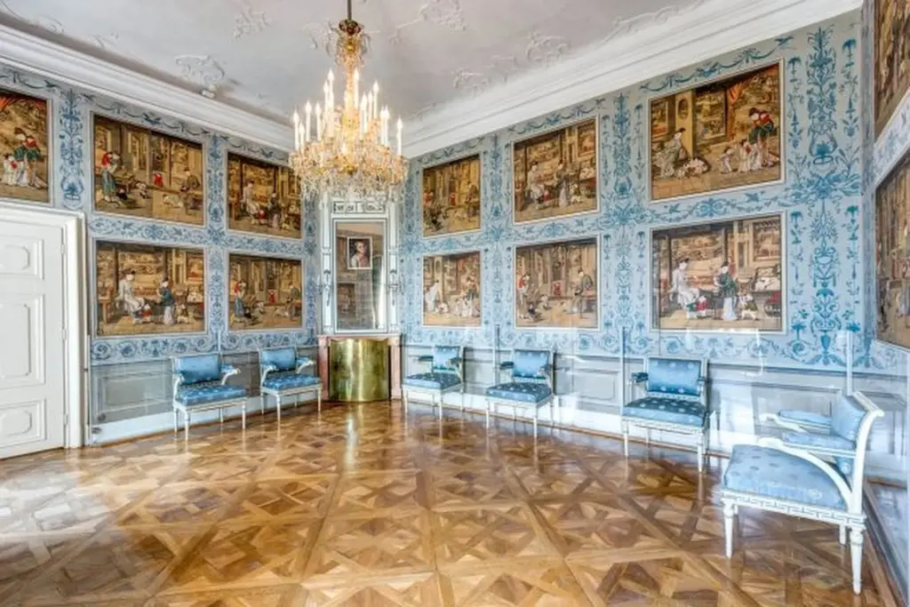 Eleganter, verschnörkelter Raum mit Parkettboden, blauen und weißen Wänden mit gerahmten Gemälden, blauen gepolsterten Stühlen an den Wänden, einem goldverzierten Tisch und einem großen Kristallkronleuchter an der Decke.
