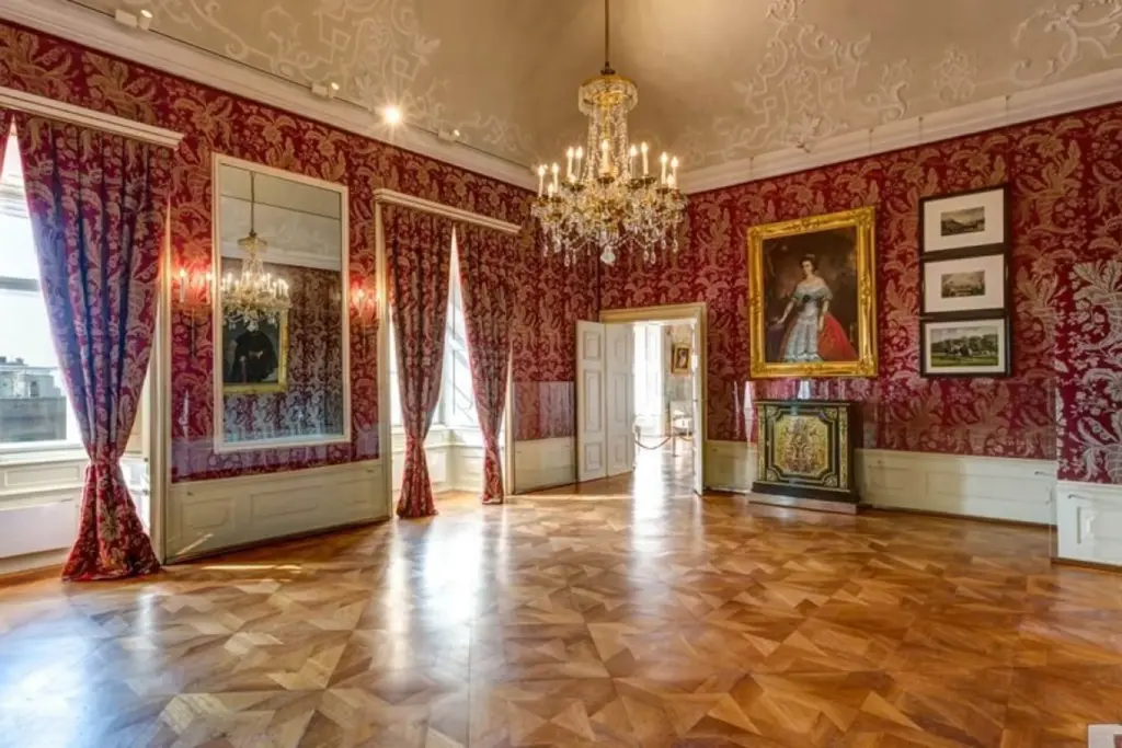 Elegantes Palastzimmer mit rot gemusterten Tapeten, großen Spiegeln, Kronleuchtern, Holzparkettboden, hohen Fenstern mit Vorhängen und gerahmten Porträts an den Wänden. Der Raum ist geräumig und üppig dekoriert.