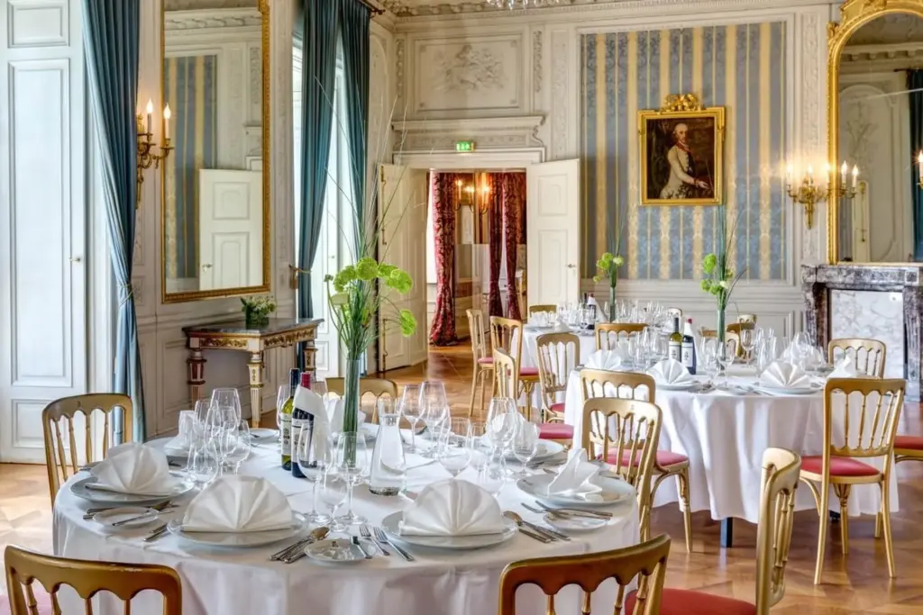 Elegantes Esszimmer mit runden Tischen, die für eine formelle Veranstaltung gedeckt sind; jeder Tisch ist mit weißen Tüchern, gefalteten Servietten, Gläsern und grünen Blumensträußen gedeckt. Der Raum verfügt über verzierte Dekoration, große Spiegel und ein Porträt an der Wand.