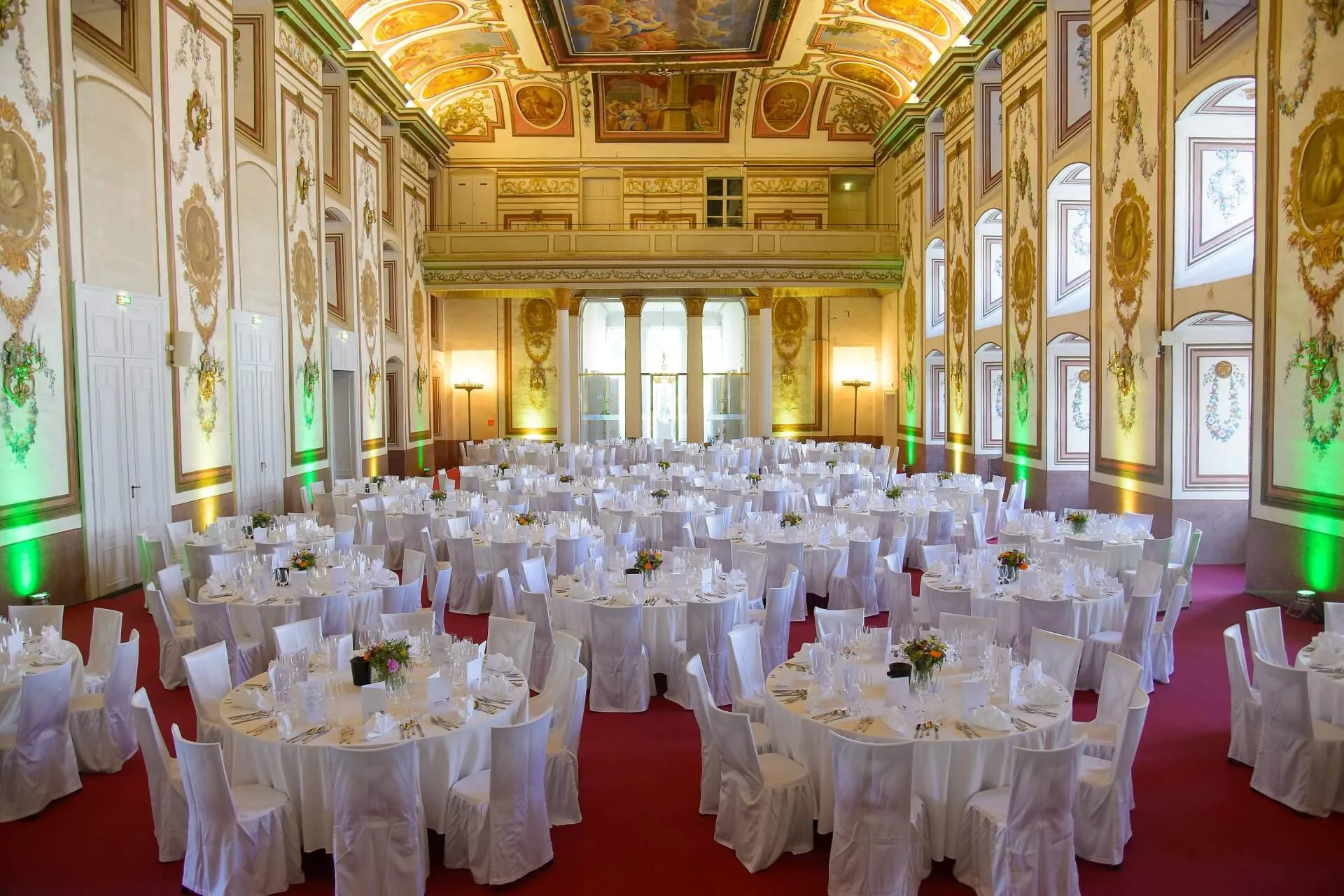 Ein großer Ballsaal mit verzierten, bemalten Decken und hohen Fenstern, eingerichtet für eine Veranstaltung mit runden Tischen, die mit weißen Tüchern und Stühlen bedeckt sind, jeder Tisch ist mit Blumen geschmückt.
