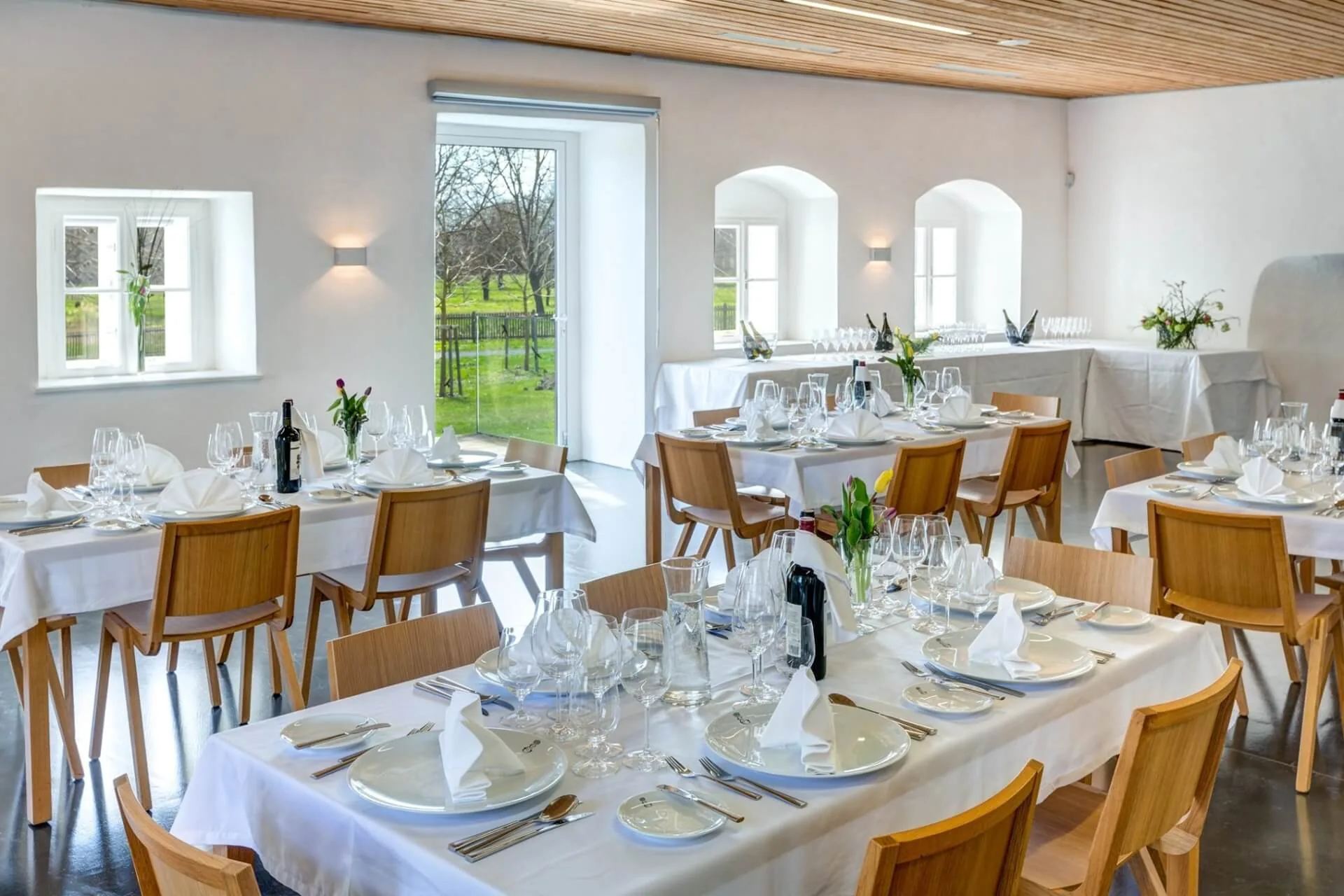 Ein heller, moderner Speisesaal mit mehreren Tischen, die für ein formelles Essen gedeckt sind, mit weißen Tischtüchern, Tellern, Gläsern, Servietten, Besteck und frischen Blumen. Große Fenster lassen viel natürliches Licht herein.
