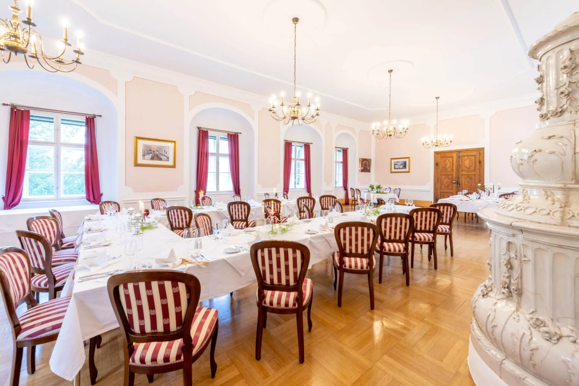 Eleganter Speisesaal mit langen Tischen, die für ein formelles Essen gedeckt sind, mit rot gestreiften Stühlen, Kronleuchtern, großen Fenstern mit roten Vorhängen, hellen Wänden und polierten Holzböden.