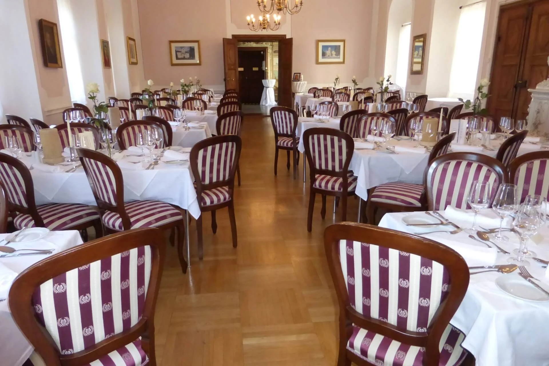 Ein formeller Speisesaal mit Holzstühlen mit rot-weiß gestreiften Polstern, weißen Tischdecken, gedeckten Tischen mit Gläsern und Besteck und von der Decke hängenden Kronleuchtern.