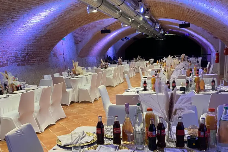 Ein eleganter Bankettsaal mit gewölbten Ziegeldecken und weiß gedeckten Tischen, die für eine Veranstaltung gedeckt sind, dekoriert mit Getränkeflaschen, Tellern, Gläsern und floralen Tafelaufsätzen unter sanfter blauer Beleuchtung.