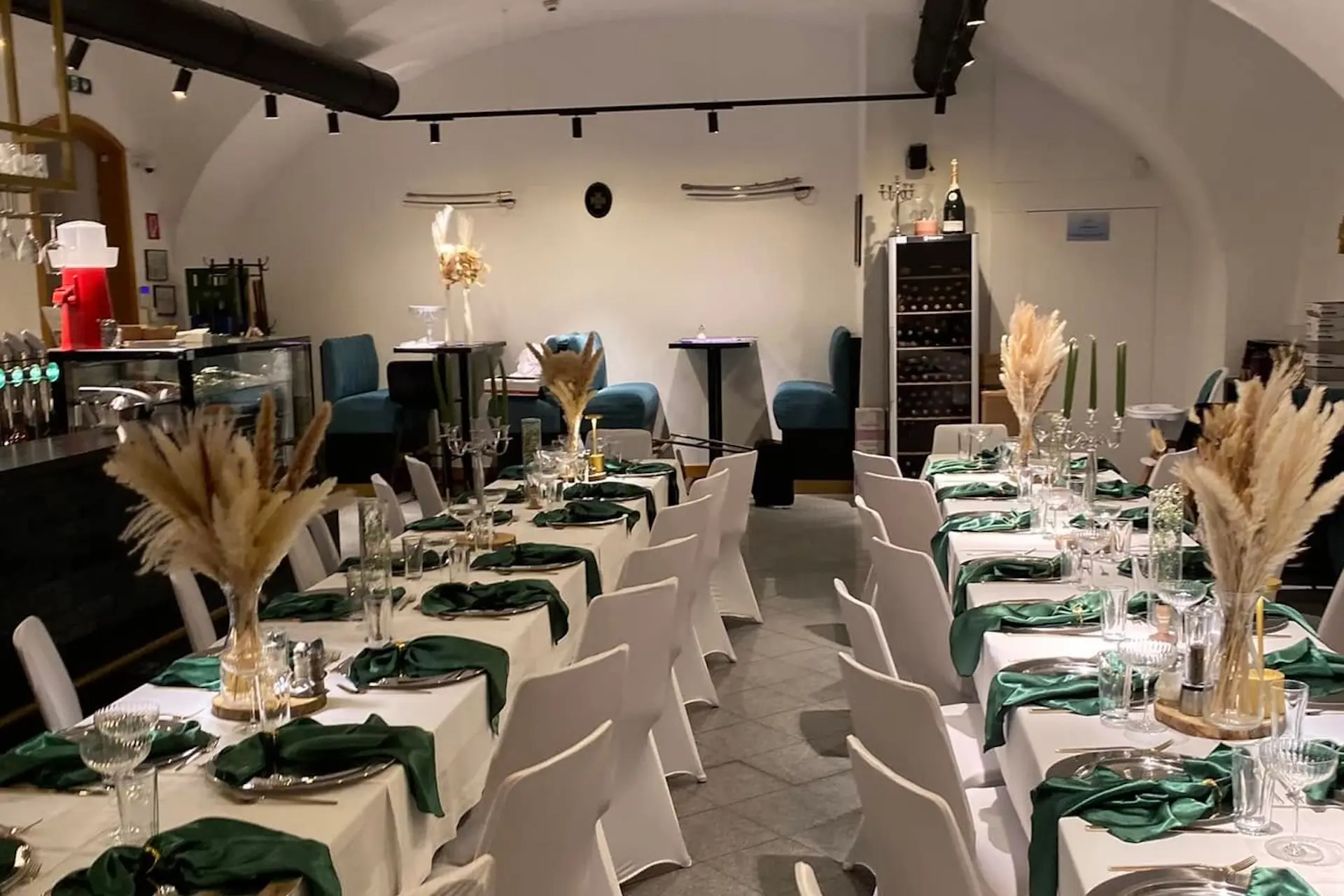 Der Essbereich eines modernen Restaurants besteht aus zwei langen Tischen, die mit grünen Servietten, Gläsern und dekorativen Weizenkörben gedeckt sind. Weiße Stühle säumen die Tische, und im Hintergrund befinden sich Sitzgelegenheiten und ein Weinregal.