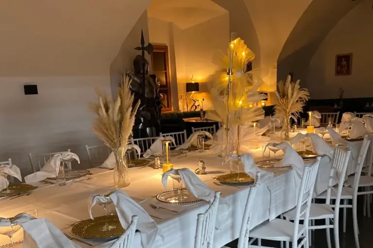 Schlosscafé Vösendorf Ein langer Esstisch ist elegant mit weißer Bettwäsche, goldumrandeten Tellern und gefalteten Servietten gedeckt und wird von warmem Kerzenlicht beleuchtet. Hohe Vasen mit Pampasgras dienen als Tafelaufsatz in einem sanft beleuchteten, gewölbten Raum.