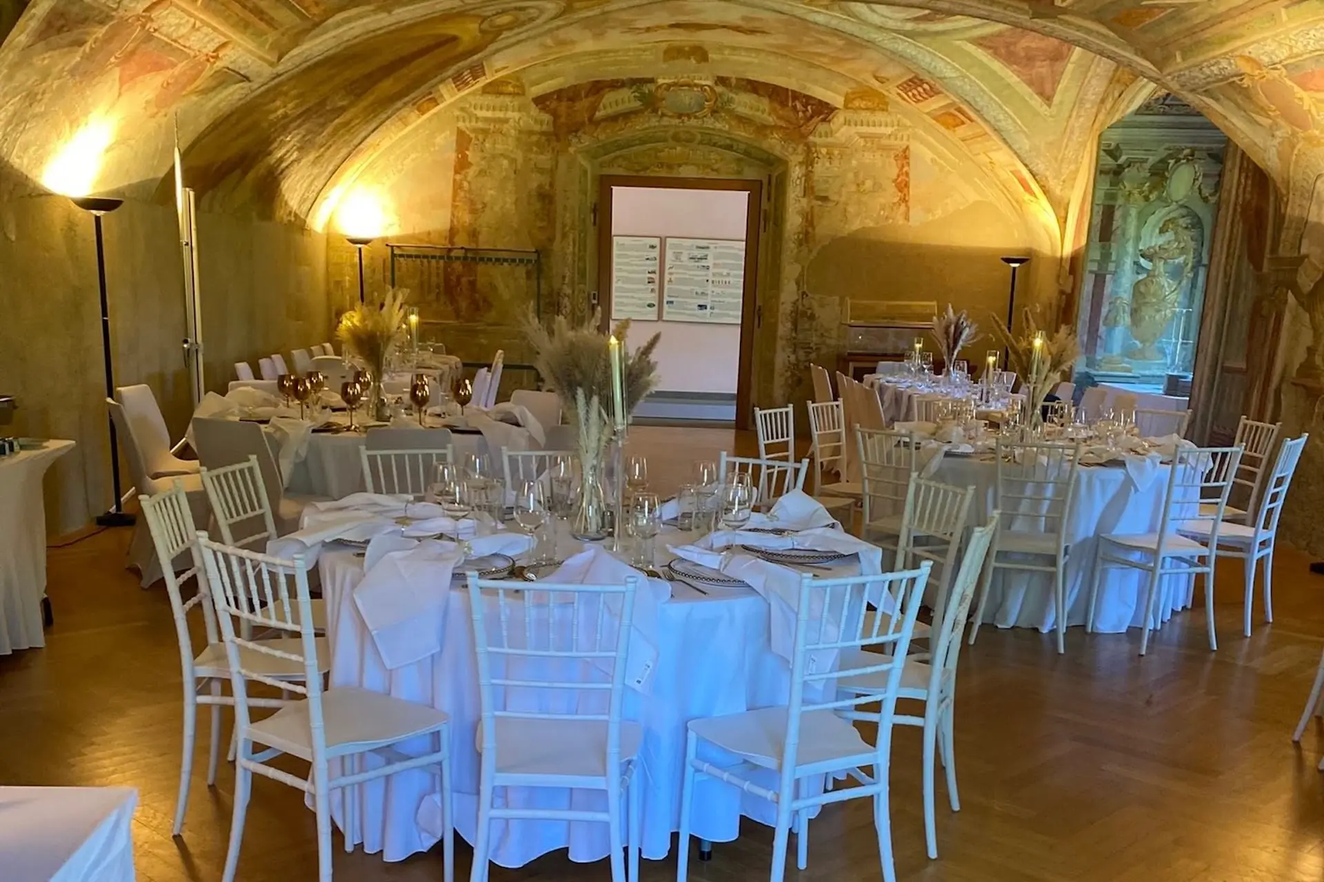 Eleganter Bankettsaal mit einer bemalten Gewölbedecke, rustikaler Wandkunst und runden Tischen mit weißen Tischdecken, Weingläsern und beigen Blumenkränzen. Weiße Stühle umgeben die Tische auf einem Holzfußboden.