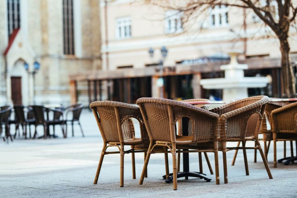 Cafétische und Korbstühle bilden einen charmanten Schanigarten auf einem ruhigen, leeren Platz mit einem Baum in der Nähe; verschwommene Gebäude und ein weißer Springbrunnen vervollständigen die heitere Szene.
