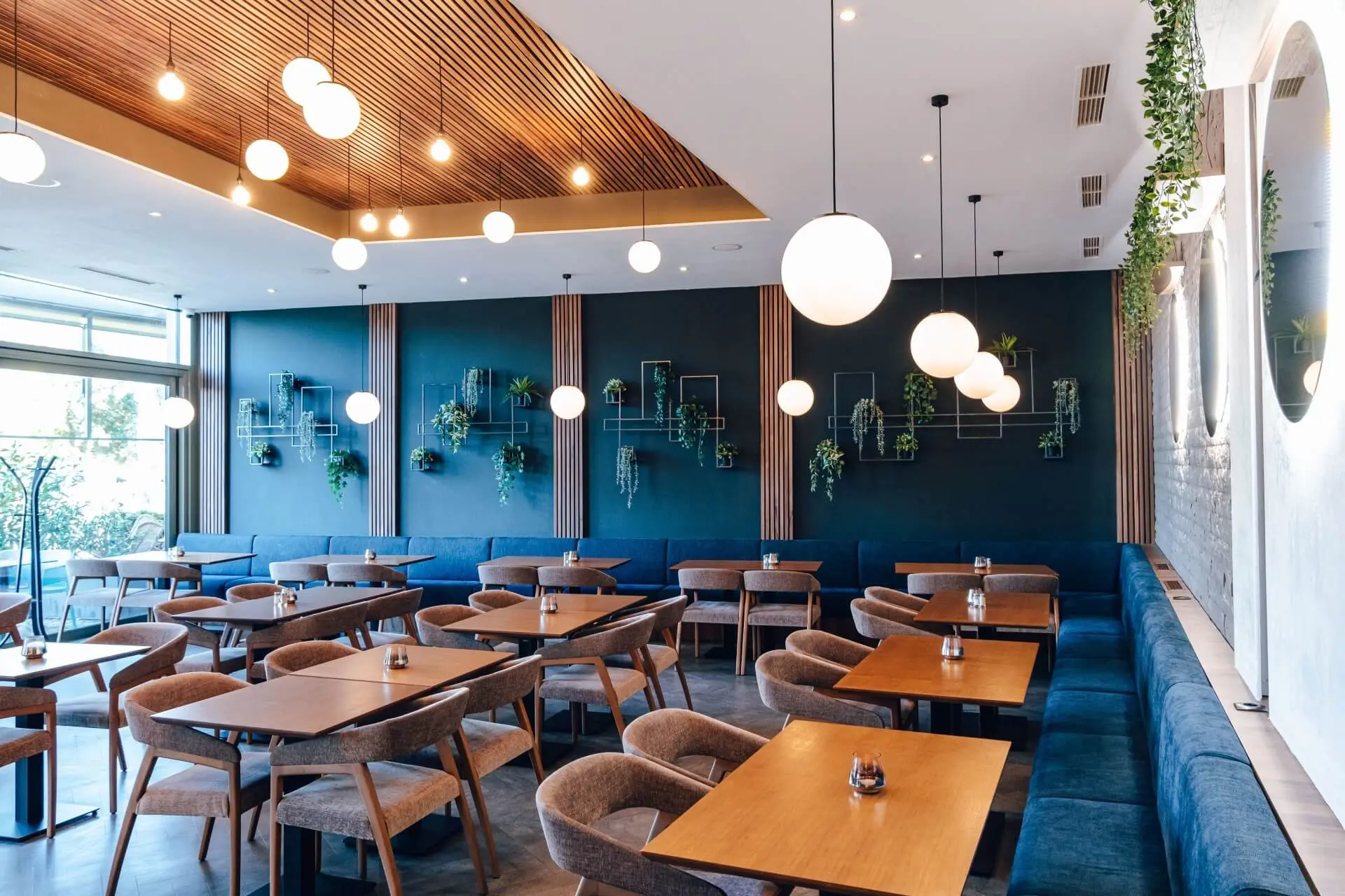 Modernes Restaurantinterieur mit Holztischen, gepolsterten Stühlen, blauer Akzentwand mit Regalen und Topfpflanzen, runden Hängelampen und einer Decke mit Holzlatten. Der Raum wirkt sauber und einladend.
