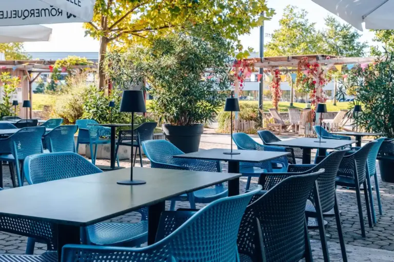 Café-Sitzbereich im Freien mit blauen Stühlen, schwarzen Tischen und kleinen schwarzen Lampen. Grüne Pflanzen und Bäume spenden Schatten, und an einem sonnigen Tag hängen bunte Dekorationen im Hintergrund.