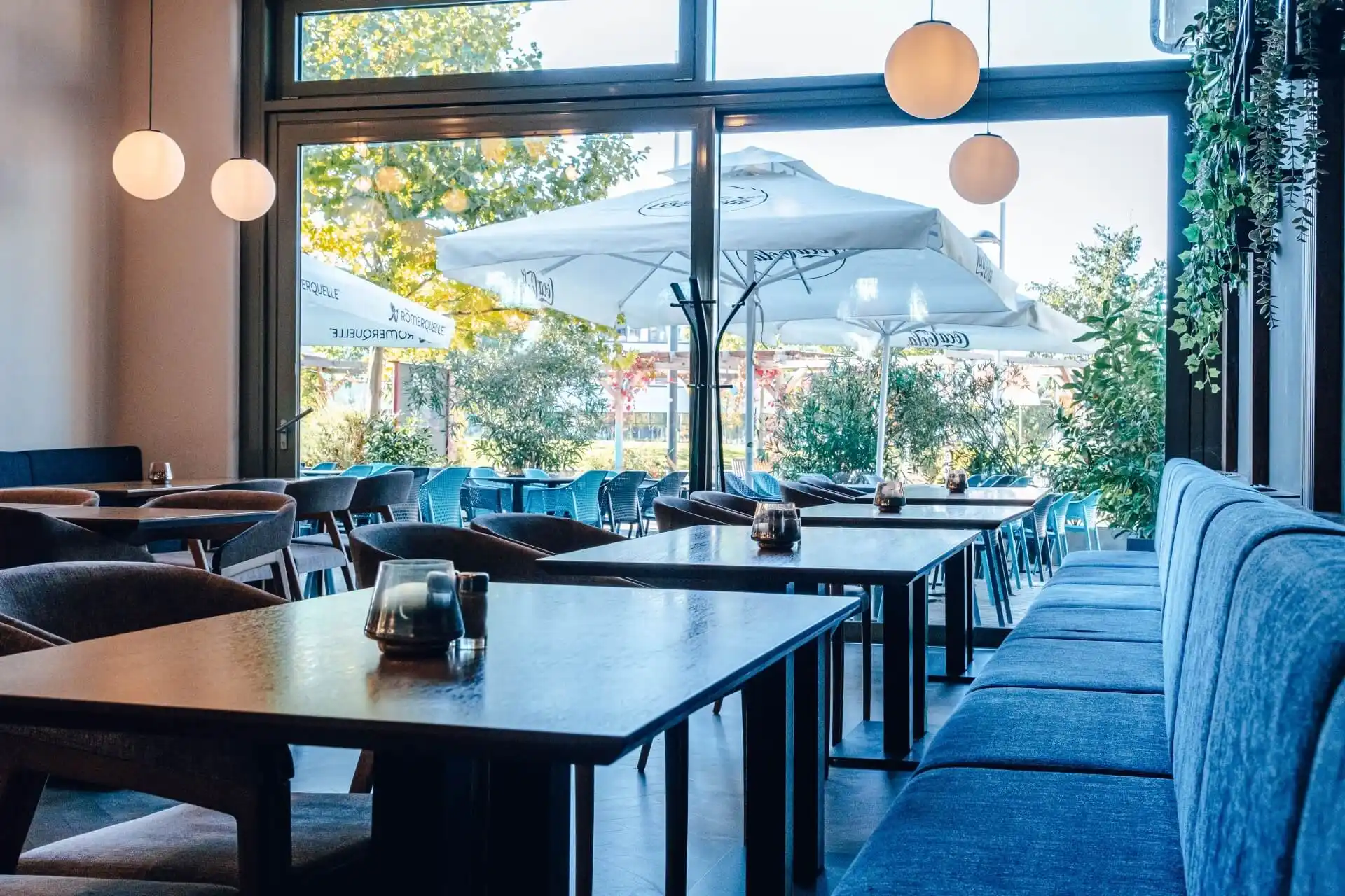 Modernes Café-Interieur mit dunklen Tischen, blau gepolsterten Sitzen und großen Fenstern, die den Blick auf eine Außenterrasse mit Sonnenschirmen, Grünpflanzen und durchdringendem Sonnenlicht freigeben. Die Atmosphäre ist hell und einladend.