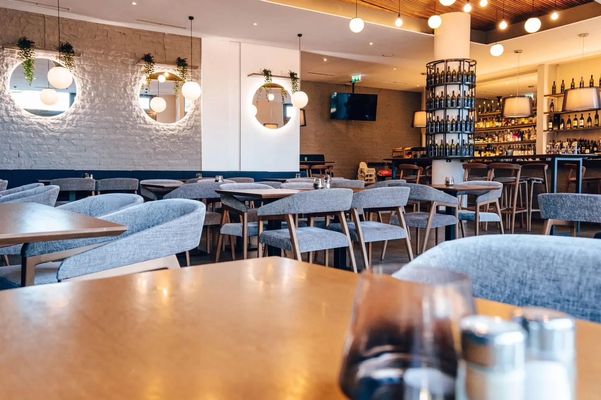 Modernes Restaurantinterieur mit Holztischen, grauen gepolsterten Stühlen, runden Wandspiegeln mit Hängepflanzen, Kugelleuchten, einem Weinregal und einem Barbereich im Hintergrund. Der Raum ist leer und gut beleuchtet.